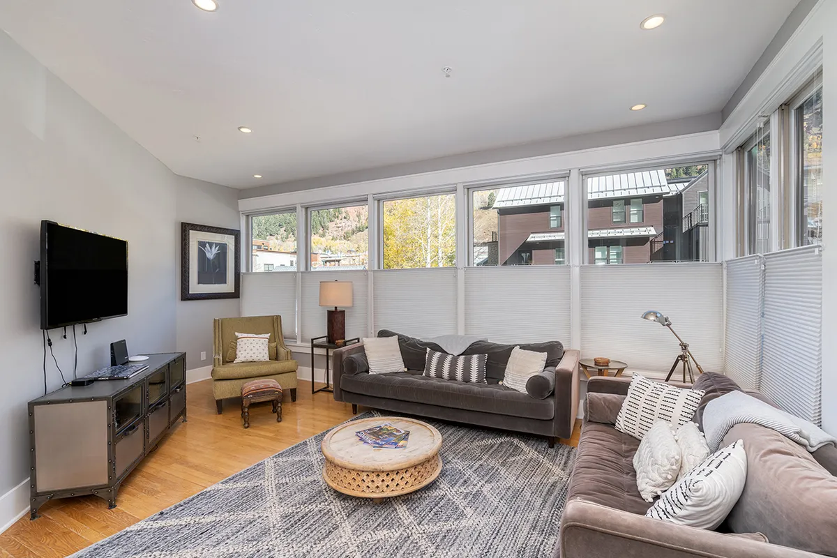 The image displays a sun-drenched living area in a Telluride condo, featuring expansive wall-to-wall windows that frame breathtaking mountain views. Cozy, stylish seating and a colorful decorative rug invite relaxation and socializing, while abundant natural light fills the open-concept space, creating a warm, welcoming atmosphere.