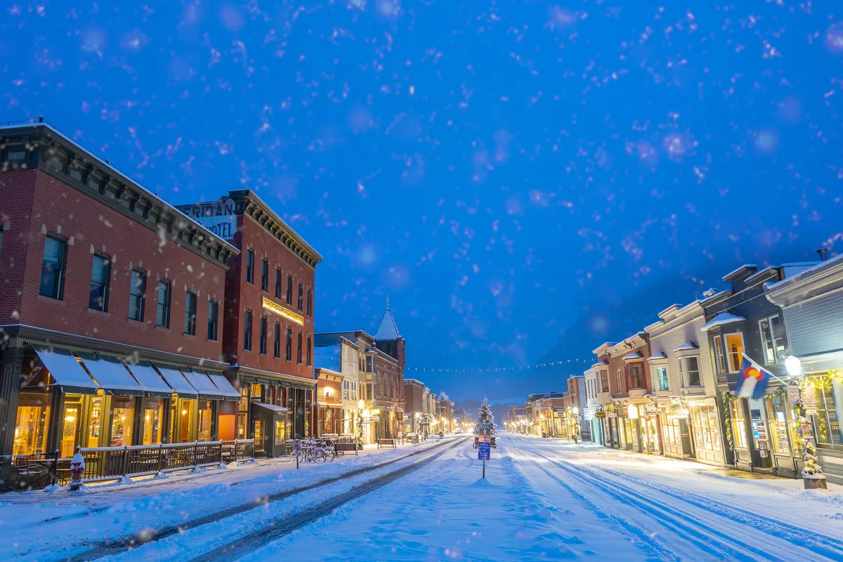 A charming winter street scene, with historic buildings, a decorated Christmas tree, warmly lit shop windows, and snow-covered streets. The atmosphere is festive and inviting, capturing the essence of Telluride’s communal holiday events and small-town vibrancy.