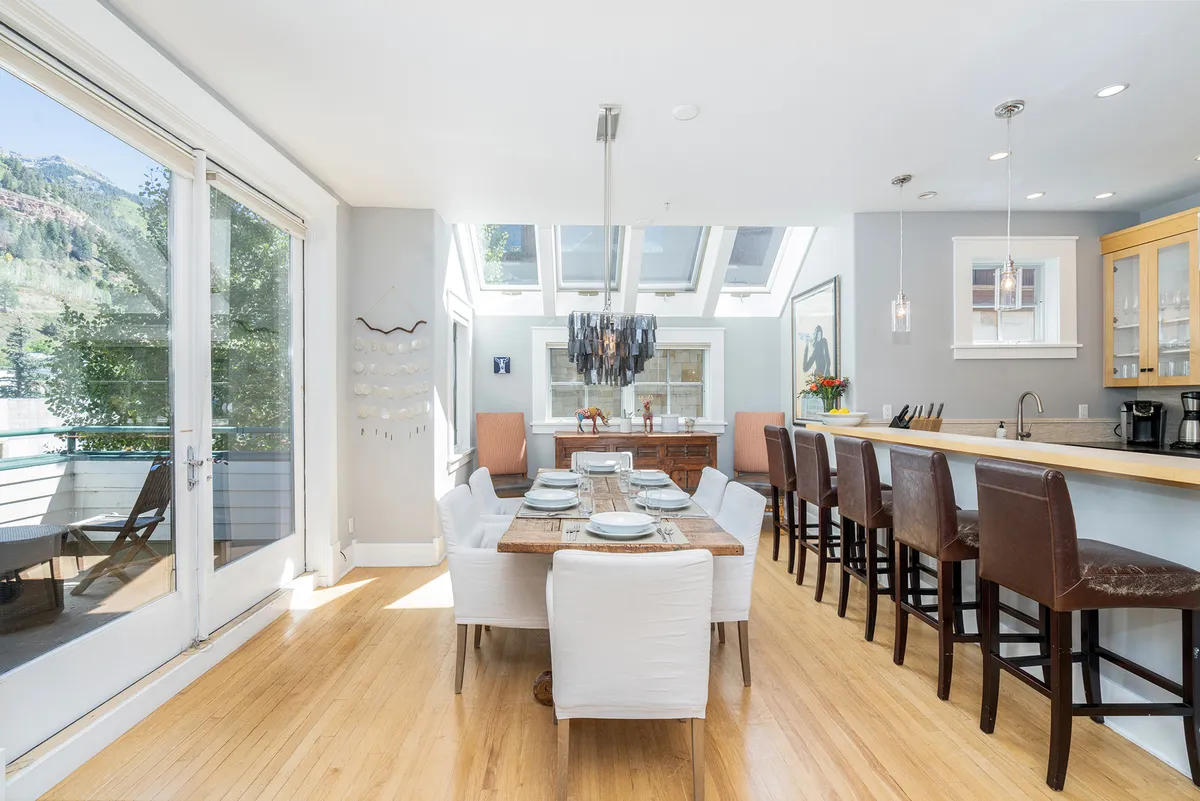 A handsomely set dining table sits beneath a stylish chandelier, adjacent to a breakfast bar. Skylights and windows fill the area with warm sunlight, and the open layout fosters a seamless flow between dining, cooking, and gathering spaces.
