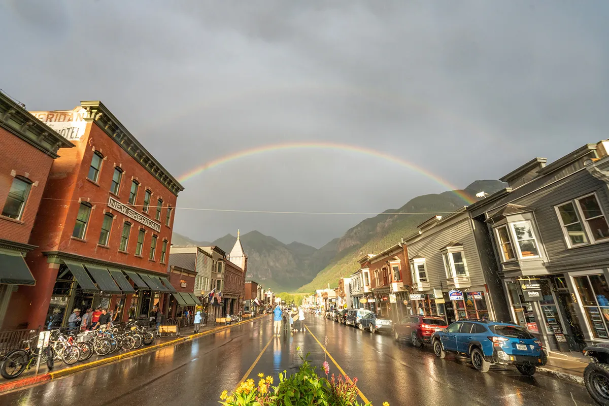 A charming Telluride street scene featuring historic buildings set against a mountain backdrop, with a rainbow arching overhead and bicycles lining the wet street. The uplifting, welcoming mood captures post-rain freshness and the town's inviting character.