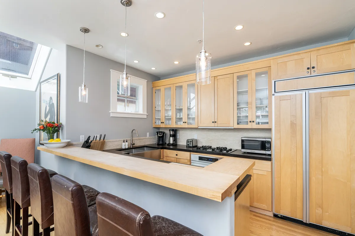 This image showcases a bright, inviting kitchen featuring a central island, stainless steel appliances, hanging light fixtures, and glass cabinet doors. The space feels open and is clearly designed for both cooking and gathering, with ample room for family or friends to connect.