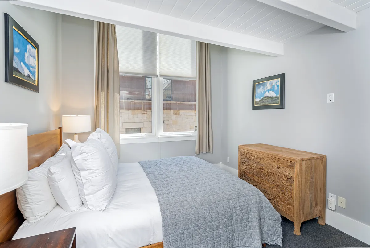 A queen bedroom with white linens, decorative pillows, a wooden nightstand with lamp, carved dresser, and artwork. The room feels bright and inviting, bathed in gentle natural light.