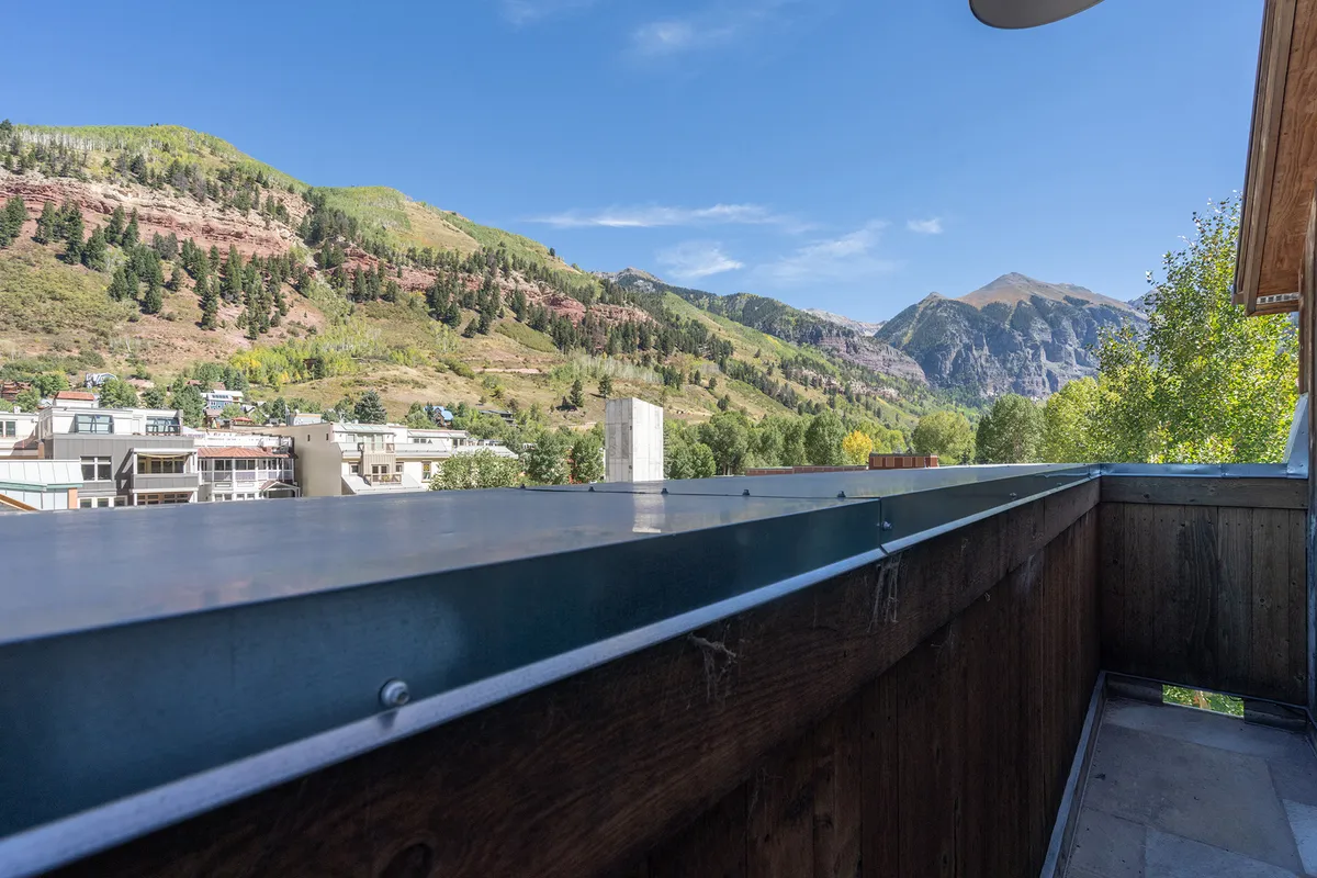 A serene mountain view as seen from a balcony or deck, with a distant blue sky and balcony railing framing the natural beauty. The tranquil setting suggests a perfect spot to relax after a day of adventure.