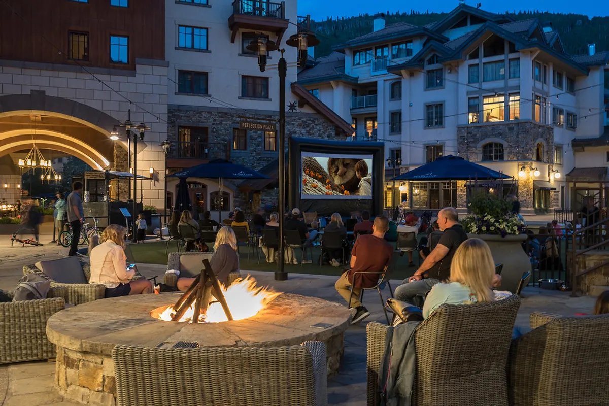 An outdoor communal space where people are gathered around a fire pit, watching a movie on an outdoor screen, with surrounding buildings in the background. The mood is relaxed and communal, and the scene evokes shared experiences and evenings spent together.