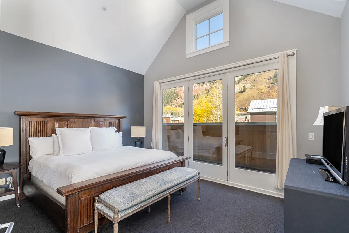 This image features a spacious bedroom with a large bed, expansive windows letting in natural light, a cozy bench, and a television. The serene atmosphere is complemented by the calming mountain vistas visible through the windows.