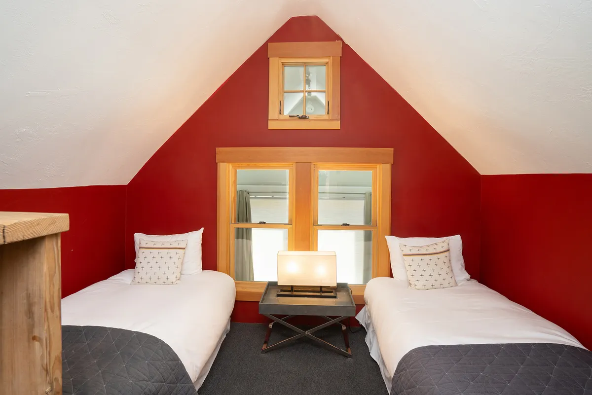 A cozy loft bedroom with two twin beds, a table lamp, and a window letting in morning light. The layout encourages shared stories and relaxation, making it ideal for kids or single travelers.